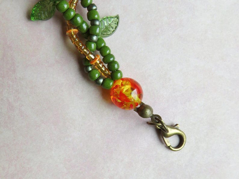 May include: A beaded keychain with a nature theme. The keychain features green beads, gold accents, and a large, round, orange and yellow glass bead. The clasp is a bronze color, and there are green leaf accents.