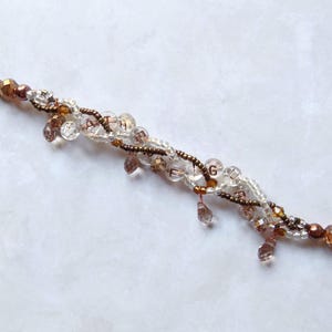 May include: A delicate bracelet featuring clear and brown beads, with a mix of round and teardrop shapes. The bracelet has a copper-colored clasp and accents, and the letters A, T, H, E, and G are visible.