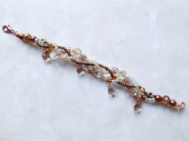 May include: A delicate bracelet featuring clear and brown beads, with a mix of round and teardrop shapes. The bracelet has a copper-colored clasp and accents, and the letters A, T, H, E, and G are visible.