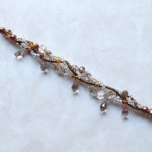 May include: A beaded bracelet with a copper clasp and accents. The bracelet features clear, brown, and gold beads, with dangling teardrop-shaped charms. The word "HEART" is spelled out with small beads. A delicate piece of jewelry.