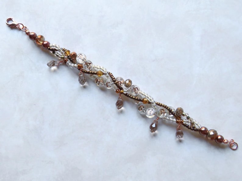 May include: A beaded bracelet with a copper clasp and accents. The bracelet features clear, brown, and gold beads, with dangling teardrop-shaped charms. The word "HEART" is spelled out with small beads. A delicate piece of jewelry.