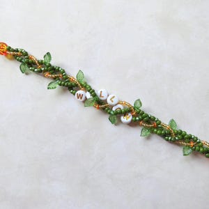 May include: A beaded bracelet with a vine-like design. The bracelet features green beads, small green leaf accents, and orange beads. White beads spell out the word "WELCOME". The bracelet is approximately 7 inches long.