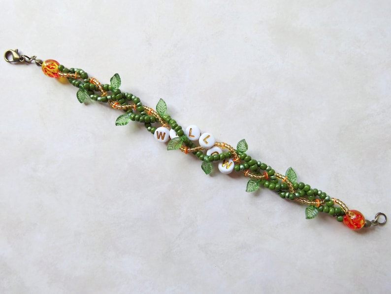 May include: A beaded bracelet with a vine-like design. The bracelet features green beads, small green leaf accents, and orange beads. White beads spell out the word "WELCOME". The bracelet is approximately 7 inches long.
