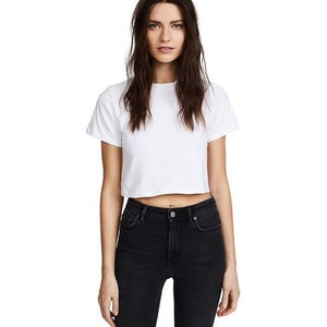 K&ouml;nnte beinhalten: Ein wei&szlig;es, kurz geschnittenes T-Shirt mit kurzen &Auml;rmeln. Dazu eine schwarze, hoch taillierte Jeans. Das Model steht im Zentrum, mit neutralem Gesichtsausdruck. Der Stil ist l&auml;ssig und minimalistisch.