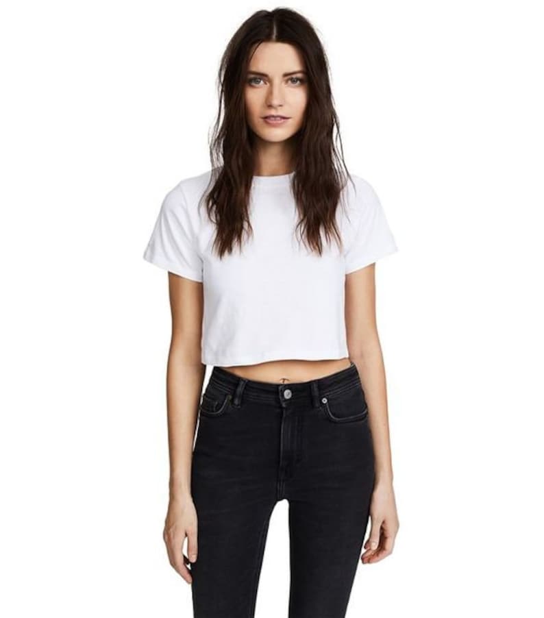K&ouml;nnte beinhalten: Ein wei&szlig;es, kurz geschnittenes T-Shirt mit kurzen &Auml;rmeln. Dazu eine schwarze, hoch taillierte Jeans. Das Model steht im Zentrum, mit neutralem Gesichtsausdruck. Der Stil ist l&auml;ssig und minimalistisch.