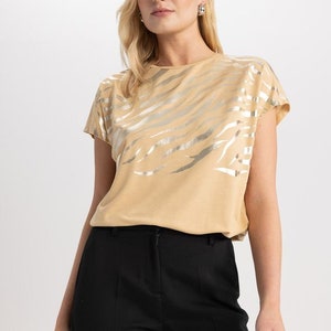 K&ouml;nnte beinhalten: Ein beiges T-Shirt mit einem silbernen Zebra-Druck. Das Shirt hat kurze &Auml;rmel und einen runden Ausschnitt.