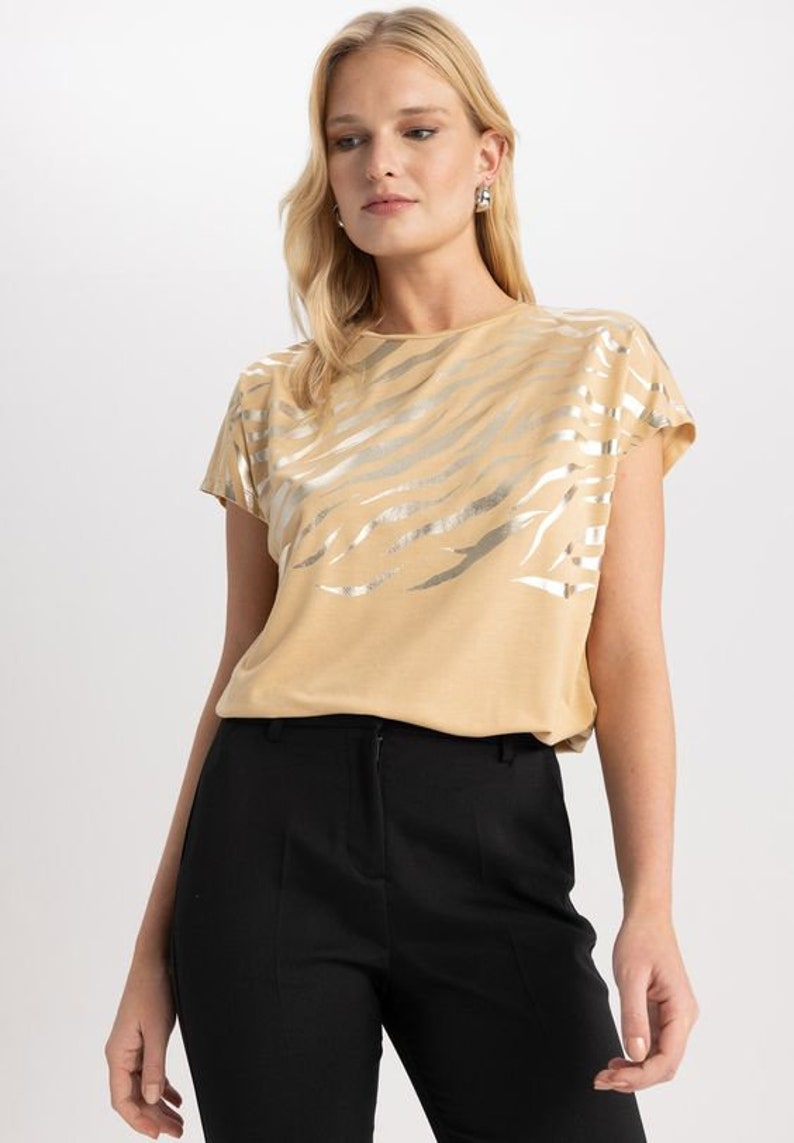 K&ouml;nnte beinhalten: Ein beiges T-Shirt mit einem silbernen Zebra-Druck. Das Shirt hat kurze &Auml;rmel und einen runden Ausschnitt.