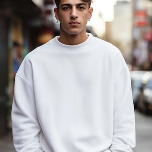 May include: A man wearing a white crew neck sweatshirt with long sleeves. He is standing in an urban setting with a blurred background.