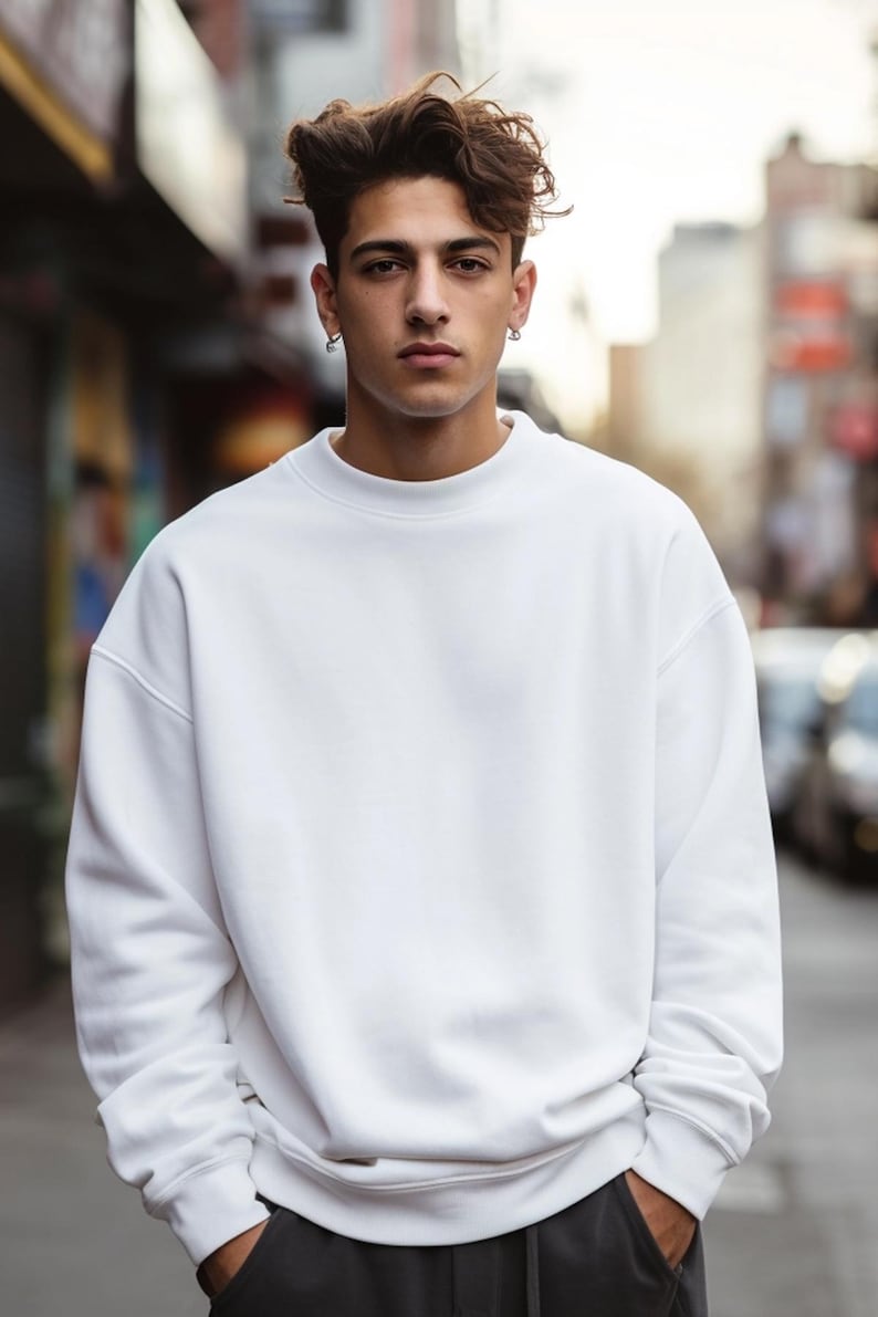 May include: A man wearing a white crew neck sweatshirt with long sleeves. He is standing in an urban setting with a blurred background.