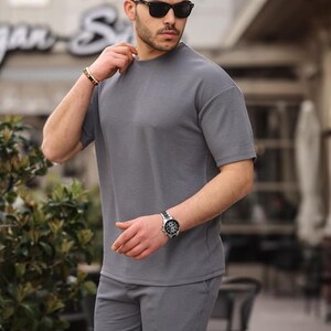 Puede incluir: Un conjunto de camiseta de manga corta y pantalones cortos a juego de color gris con textura de canal&eacute;. El modelo lleva gafas de sol y un reloj.
