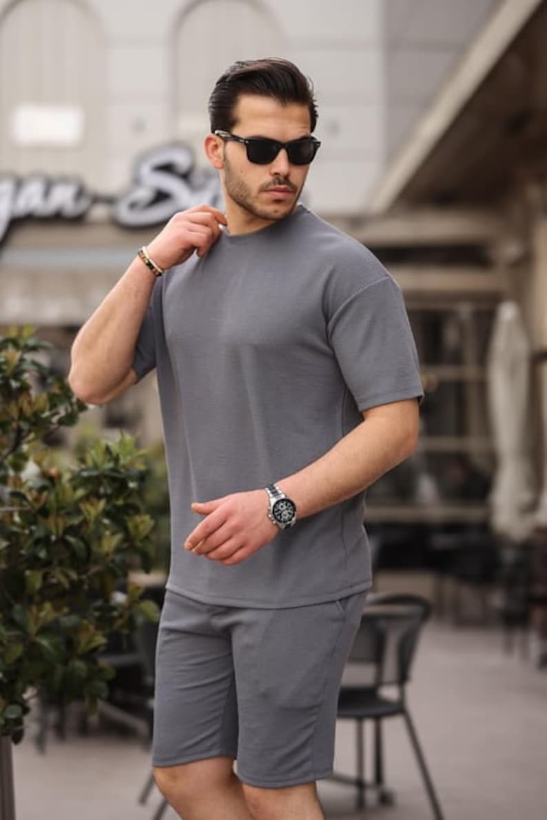 Puede incluir: Un conjunto de camiseta de manga corta y pantalones cortos a juego de color gris con textura de canal&eacute;. El modelo lleva gafas de sol y un reloj.