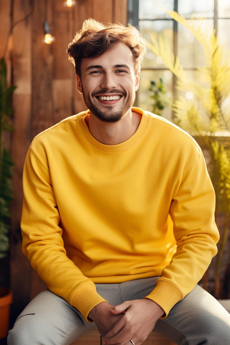 May include: A man wearing a yellow sweatshirt and grey trousers smiles at the camera.