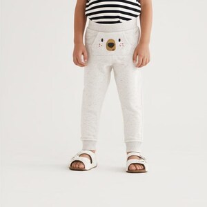 May include: A child wearing light gray sweatpants with a koala face design on the front pocket. The child is also wearing a black and white striped t-shirt and white sandals with brown soles.