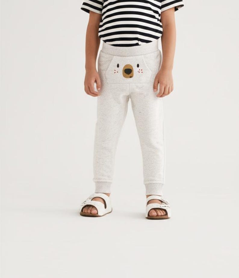 May include: A child wearing light gray sweatpants with a koala face design on the front pocket. The child is also wearing a black and white striped t-shirt and white sandals with brown soles.