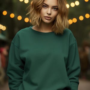 May include: A woman wearing a dark green sweatshirt with long sleeves and a crew neck. She is standing in front of a blurred background of lights.