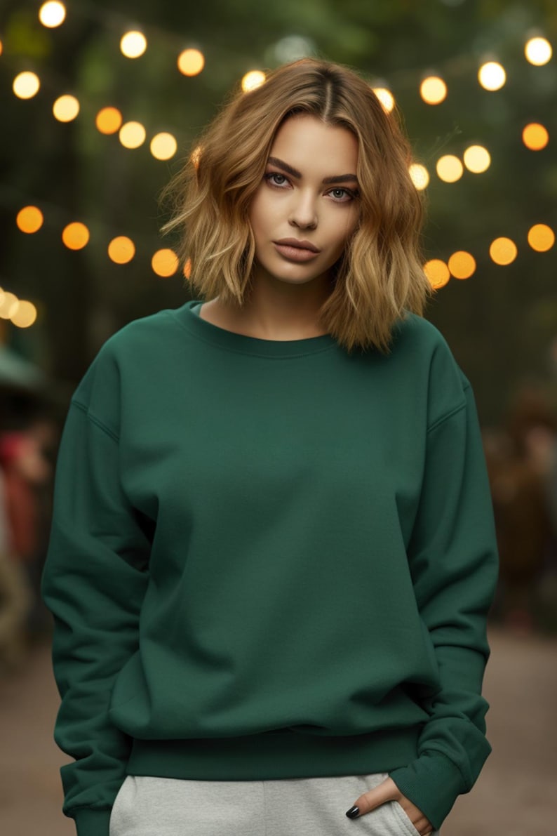 May include: A woman wearing a dark green sweatshirt with long sleeves and a crew neck. She is standing in front of a blurred background of lights.