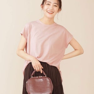 K&ouml;nnte beinhalten: Eine Frau tr&auml;gt ein rosa T-Shirt und einen braunen Rock und h&auml;lt eine braune Handtasche aus Krokodilleder mit einem oberen Griff.