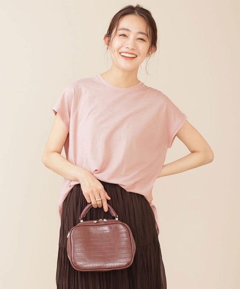 K&ouml;nnte beinhalten: Eine Frau tr&auml;gt ein rosa T-Shirt und einen braunen Rock und h&auml;lt eine braune Handtasche aus Krokodilleder mit einem oberen Griff.