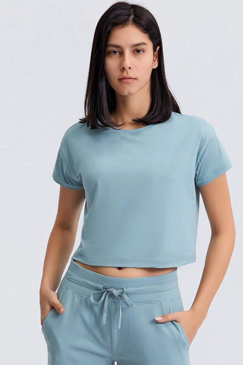 K&ouml;nnte beinhalten: Ein hellblaues Crop-Top und eine passende Jogginghose. Das kurz&auml;rmelige Oberteil hat einen Rundhalsausschnitt und einen verk&uuml;rzten Saum. Die Jogginghose hat einen Kordelzug in der Taille und Taschen. Das Model tr&auml;gt das Sportbekleidungsset.