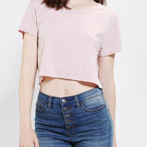 K&ouml;nnte beinhalten: Ein hellrosa Crop-Top mit kurzen &Auml;rmeln. Das Oberteil wird mit einer blauen Jeans mit Knopfleiste getragen. Die Jeans hat ein klassisches F&uuml;nf-Taschen-Design.