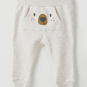 May include: Light gray baby sweatpants with a bear face design on the pocket. The bear face has brown eyes, a brown nose, and red dots for cheeks. The sweatpants have a ribbed waistband and cuffs.