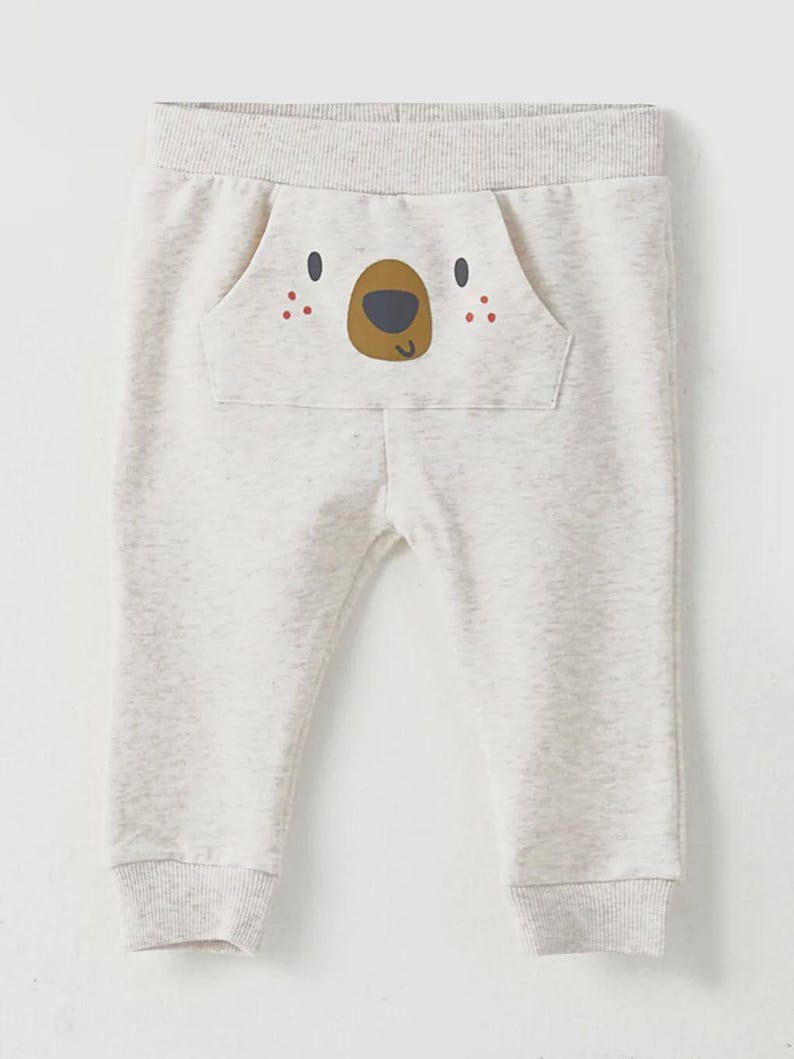 May include: Light gray baby sweatpants with a bear face design on the pocket. The bear face has brown eyes, a brown nose, and red dots for cheeks. The sweatpants have a ribbed waistband and cuffs.