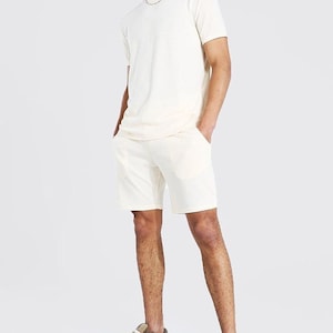 Puede incluir: Un conjunto de camiseta de manga corta y pantalones cortos a juego de color crema. El modelo lleva el conjunto con un par de zapatillas de deporte estampadas.