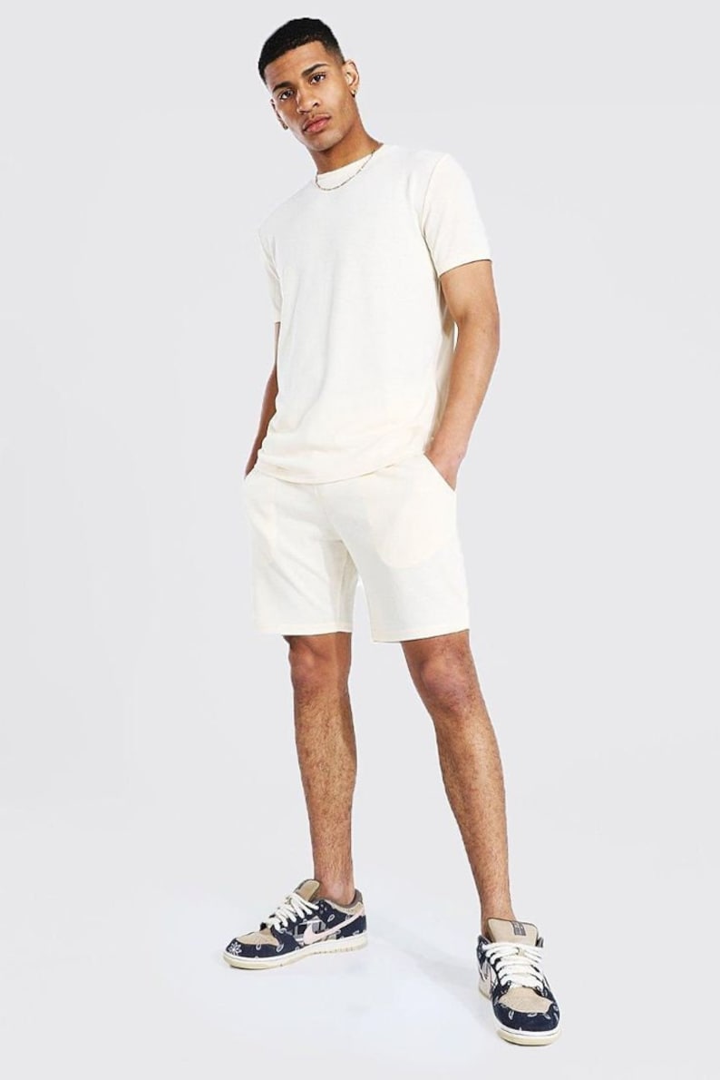 Puede incluir: Un conjunto de camiseta de manga corta y pantalones cortos a juego de color crema. El modelo lleva el conjunto con un par de zapatillas de deporte estampadas.