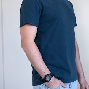 May include: A man wearing a dark blue t-shirt and light blue jeans. He is wearing a black digital watch on his left wrist.