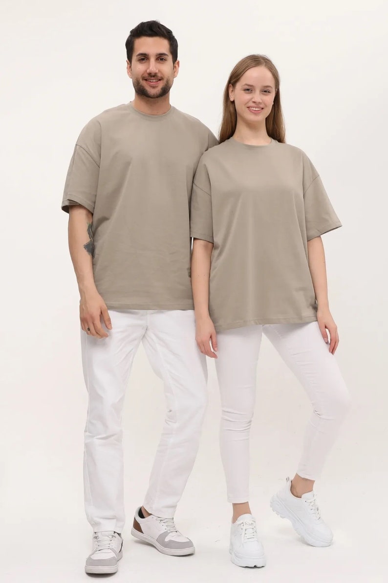 Puede incluir: Dos personas con camisetas de color marr&oacute;n claro de gran tama&ntilde;o y pantalones y zapatos blancos. Las camisetas tienen cuello redondo y mangas cortas.