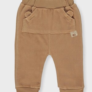 May include: Tan baby pants with a large front pocket and small ear-shaped details. The pants have a ribbed waistband and cuffs. The fabric appears soft and comfortable. A small tag is sewn on the pocket. These pants are suitable for infants and toddlers.