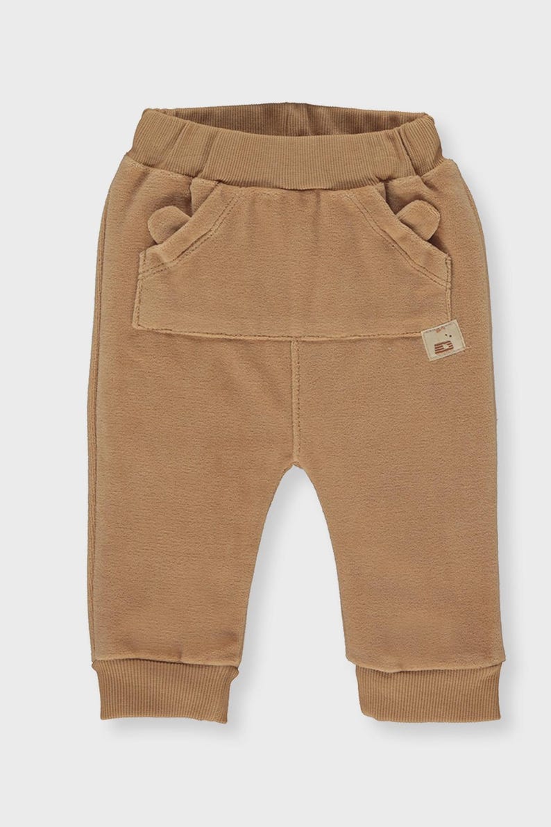 May include: Tan baby pants with a large front pocket and small ear-shaped details. The pants have a ribbed waistband and cuffs. The fabric appears soft and comfortable. A small tag is sewn on the pocket. These pants are suitable for infants and toddlers.
