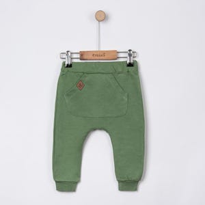 May include: Olive green baby pants with a large front pocket and a small brown patch. The pants are hanging on a wooden hanger with the brand name "tecixs" printed on it. The pants have a relaxed fit and cuffed ankles.