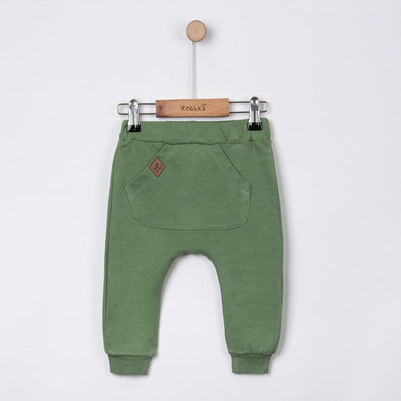 May include: Olive green baby pants with a large front pocket and a small brown patch. The pants are hanging on a wooden hanger with the brand name "tecixs" printed on it. The pants have a relaxed fit and cuffed ankles.