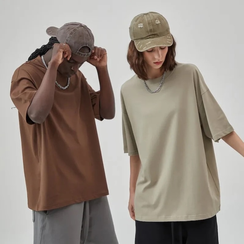 Puede incluir: Dos personas con camisetas de gran tama&ntilde;o. Una persona lleva una camiseta marr&oacute;n y una gorra de b&eacute;isbol marr&oacute;n. La otra persona lleva una camiseta verde claro y una gorra de b&eacute;isbol verde claro.