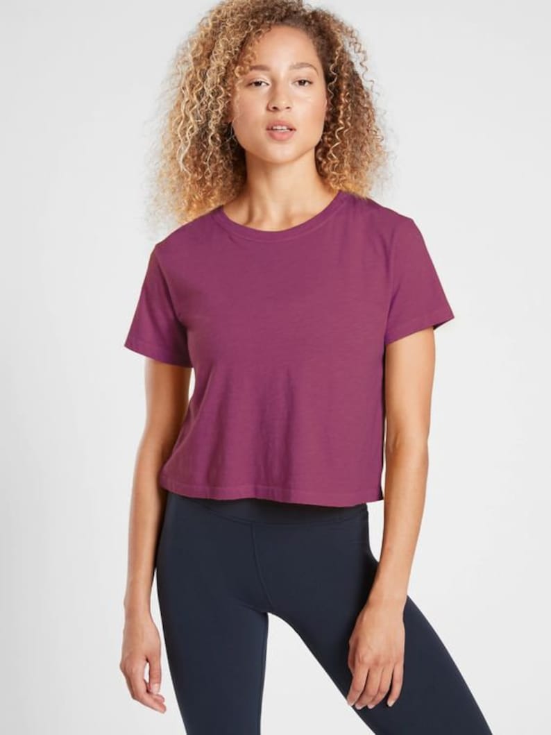 K&ouml;nnte beinhalten: Ein kurz&auml;rmeliges, bauchfreies T-Shirt in einem tiefen Magenta-Farbton. Das Shirt hat einen Rundhalsausschnitt und eine l&auml;ssige Passform. Kombiniert mit dunkelblauen Leggings ist dieses Outfit f&uuml;r sportliche Aktivit&auml;ten oder den Alltag geeignet.