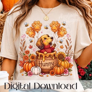 Könnte beinhalten: Ein beiges T-Shirt mit einer Grafik eines braunen Dackels in einem roten Pullover, der in einem mit Hunny beschrifteten Honigtopf sitzt. Das Design zeigt orangefarbene Kürbisse, Sonnenblumen, Herbstblätter, Bienen und pfotenförmige Luftballons.
