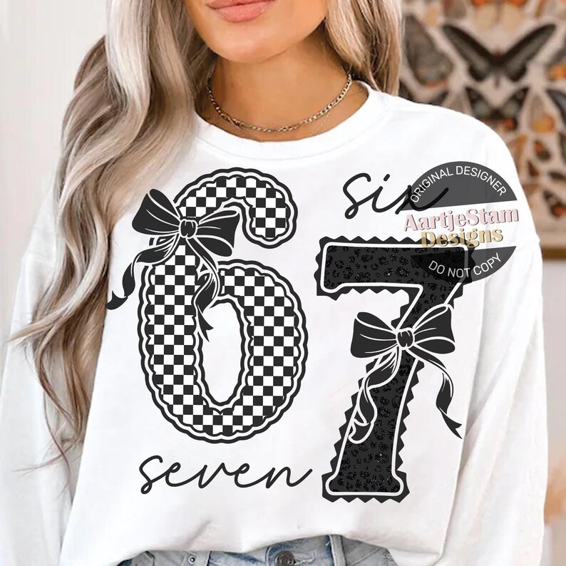 May include: White sweatshirt featuring a black and white checkered number six with a bow, and a black number seven with a bow and leopard print. The word "seven" is written in script below the numbers.
