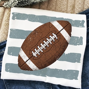 Könnte beinhalten: Ein weißes T-Shirt mit einer großen, braunen Grafik eines American Footballs mit Used-Optik, zentriert über horizontalen, verblassten blaugrauen Pinselstrich-Streifen. Der Football hat weiße Schnürsenkel und weiße Streifen an den Enden im Vintage-Stil.