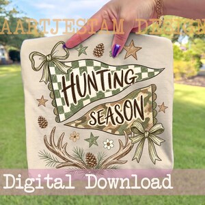 Könnte beinhalten: Ein beiges T-Shirt mit einem Grafikdesign aus zwei Wimpeln, die den Text "HUNTING SEASON" zeigen. Das Design umfasst grüne Karomuster und Tarnmuster, braune Geweihe, Tannenzapfen, Sterne, Blumen und zwei hellgrüne Schleifen.