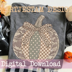 Könnte beinhalten: Ein anthrazitfarbenes T-Shirt mit einer großen Kürbisgrafik und einer Schleife mit Leopardenmuster. Der Kürbis hat abwechselnde Abschnitte mit Camouflage, beigefarbenem Karomuster und salbeigrün mit weißen Punkten. Text: ARTJESTAM DESIGN.