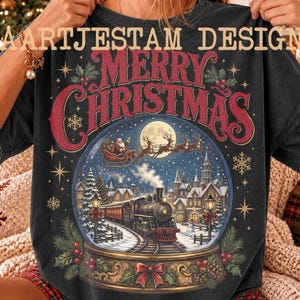 Könnte beinhalten: Ein anthrazitfarbenes T-Shirt mit einer großen Grafik einer Schneekugel, die einen Dampfzug, ein Dorf und den Weihnachtsmann im Schlitten zeigt. Der Text MERRY CHRISTMAS steht in großen roten Buchstaben darüber, mit AARTJESTAM DESIGN.