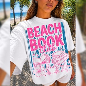 Könnte beinhalten: Ein weißes T-Shirt mit kurzen Ärmeln und einem Grafikdruck mit dem Text BEACH BOOK club in fetten rosa Buchstaben. Das Design enthält blaue vertikale Streifen, Illustrationen von offenen und gestapelten Büchern, eine Hibiskusblüte, eine Krabbe und einen Seestern.