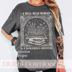 Könnte beinhalten: Ein anthrazitfarbenes Kurzarm-T-Shirt mit einem weißen Grafikdruck eines offenen Buches, aus dessen Seiten Blumen wachsen, umgeben von einem verzierten Rahmen, einer Mondsichel und dem Text A WELL-READ WOMAN IS A DANGEROUS CREATURE.