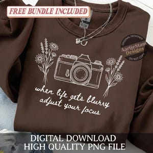 Könnte beinhalten: Ein braunes Sweatshirt mit einer weißen Grafik einer Kamera, Blumen und dem Text "when life gets blurry adjust your focus". Der Text "Free Bundle Included" und "Digital Download High Quality PNG File" ist ebenfalls sichtbar.