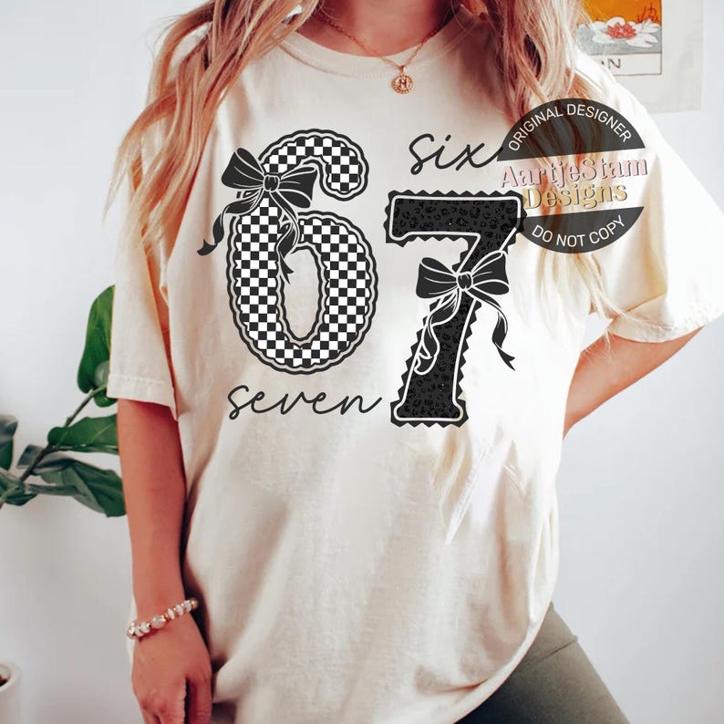 May include: Cream-colored t-shirt featuring the numbers "67" with bow accents. The number "6" has a black and white checkered pattern, while the "7" has a black leopard print. The word "seven" is written below the number "6".