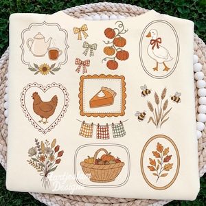 May include: A cream-colored t-shirt featuring a collection of autumn-themed illustrations including a teapot, a goose with a bow, a chicken in a heart frame, a slice of pumpkin pie, bees, wheat, a basket of pumpkins, and various plaid bows.
