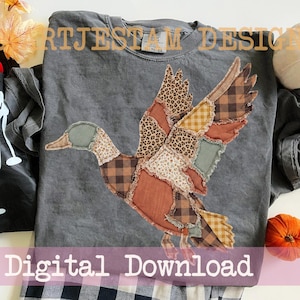 Könnte beinhalten: Ein anthrazitfarbenes T-Shirt mit einer großen, zentralen Applikation einer fliegenden Ente. Die Ente besteht aus verschiedenen Stoffstücken mit Mustern wie Leopardenprint, braunem Karomuster, gelbem Vichy-Karo sowie einfarbigem Rost und Salbeigrün.