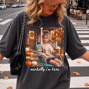 Könnte beinhalten: Ein anthrazitfarbenes Oversize-T-Shirt mit einem Grafikdruck einer Puppe, die auf einem türkisfarbenen Stuhl auf einer Veranda mit Kürbissen sitzt und einen Kaffee hält. Der Text "mentally i'm here" steht in weißer Schrift darunter.