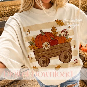Könnte beinhalten: Ein cremefarbenes T-Shirt mit einer Grafik eines hölzernen Wagens, der mit orangefarbenen Kürbissen, einem roten Apfel und einem weißen Gänseblümchen gefüllt ist. Das Design enthält Herbstblätter, Beeren und kleine Sterne sowie den Text Aartjestam Designs.